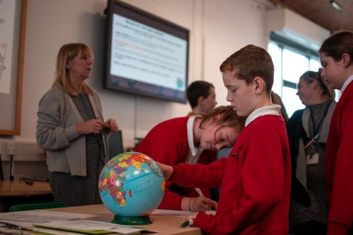 Young pupils in school uniforms standing around a table, busy at work, One pupil looking at a globe. An adult standing in the background with her back to a television screen