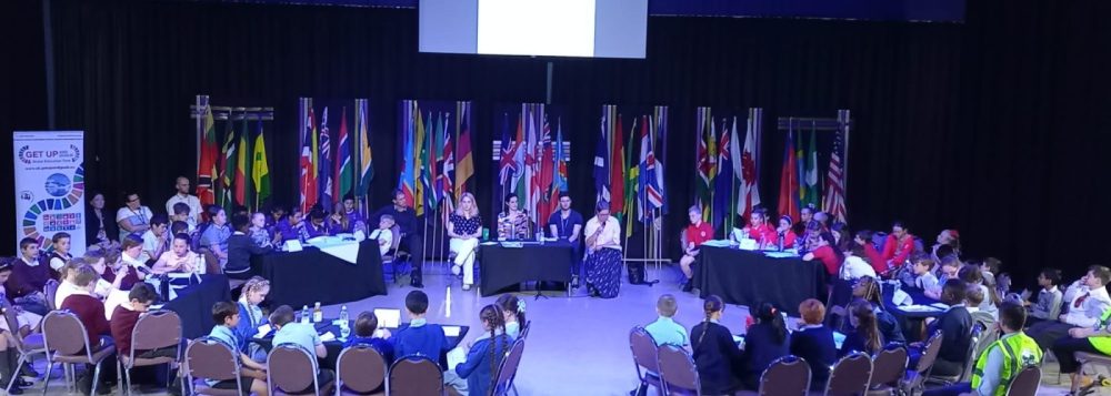 A room full of adult panelists and primary school participants at a climate conference sitting in a circle format around a large room, in the backdrop flags of several member states of the United Nations